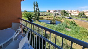 A balcony with white chairs and a view of a body of water, greenery, and buildings in the distance. - Apartment 'Cap d'Agde - Primavera - 2' with Sea View, Shared Pool and Air Conditioning (Agde)