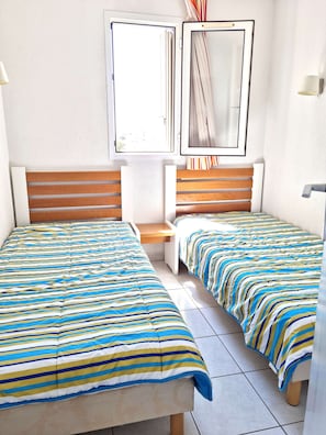 A room with two beds, a window, a bedside table, and a lamp. - Apartment 'Cap d'Agde - Primavera - 2' with Sea View, Shared Pool and Air Conditioning (Agde)