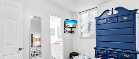 Une chambre à coucher comprenant un lit, une commode, un téléviseur et une fenêtre avec des stores.