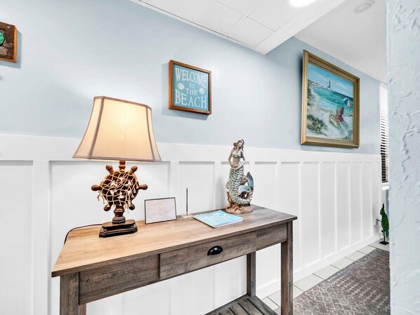 A wooden desk with a lamp, a framed welcome sign, a mermaid statue, and a framed painting of a beach scene. - Sunbird 212e (Panama City Beach)