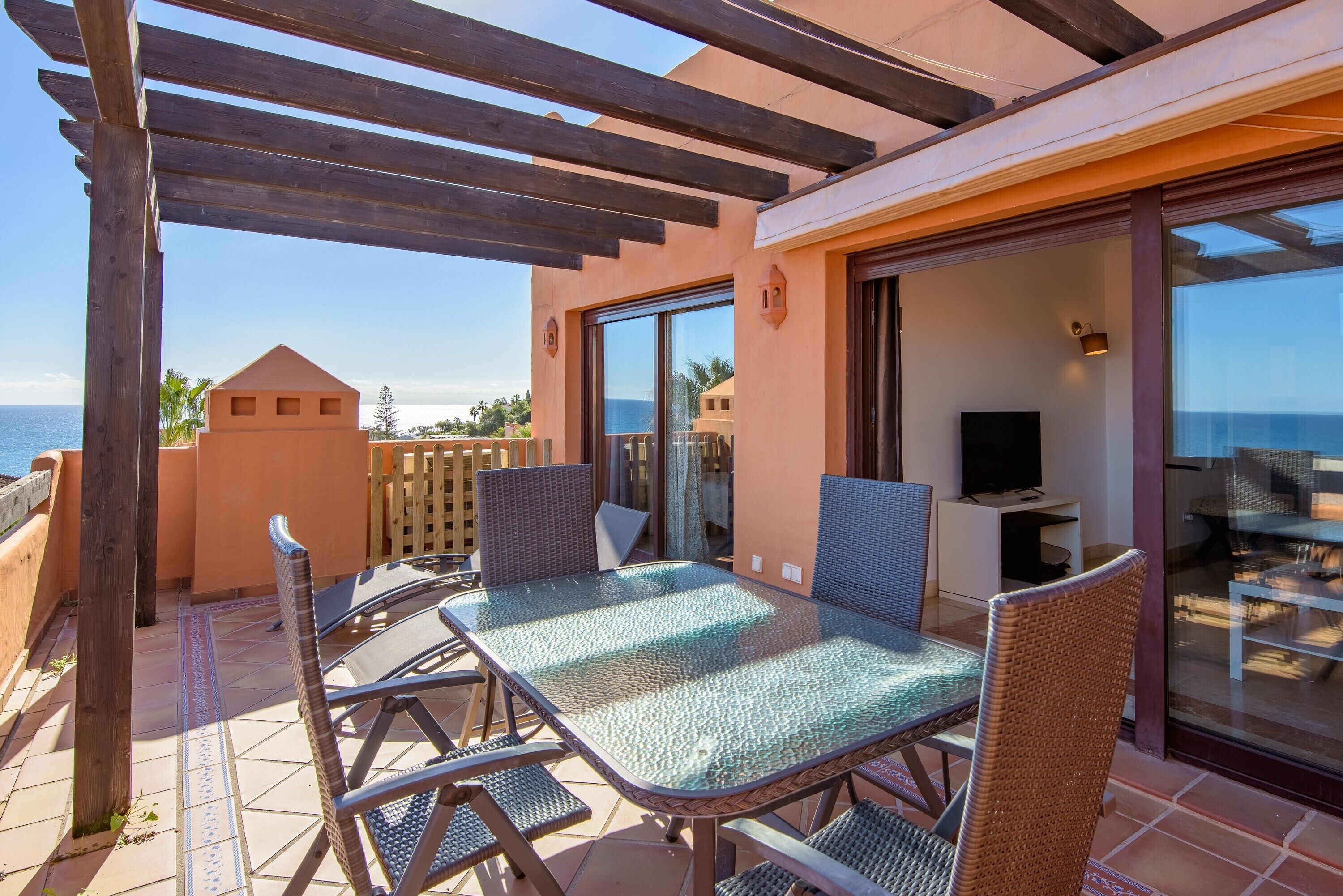 A covered patio with a glass table and wicker chairs, a TV, and a view of the sea.