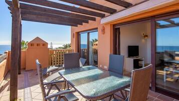 A covered patio with a glass table and wicker chairs, a TV, and a view of the sea.