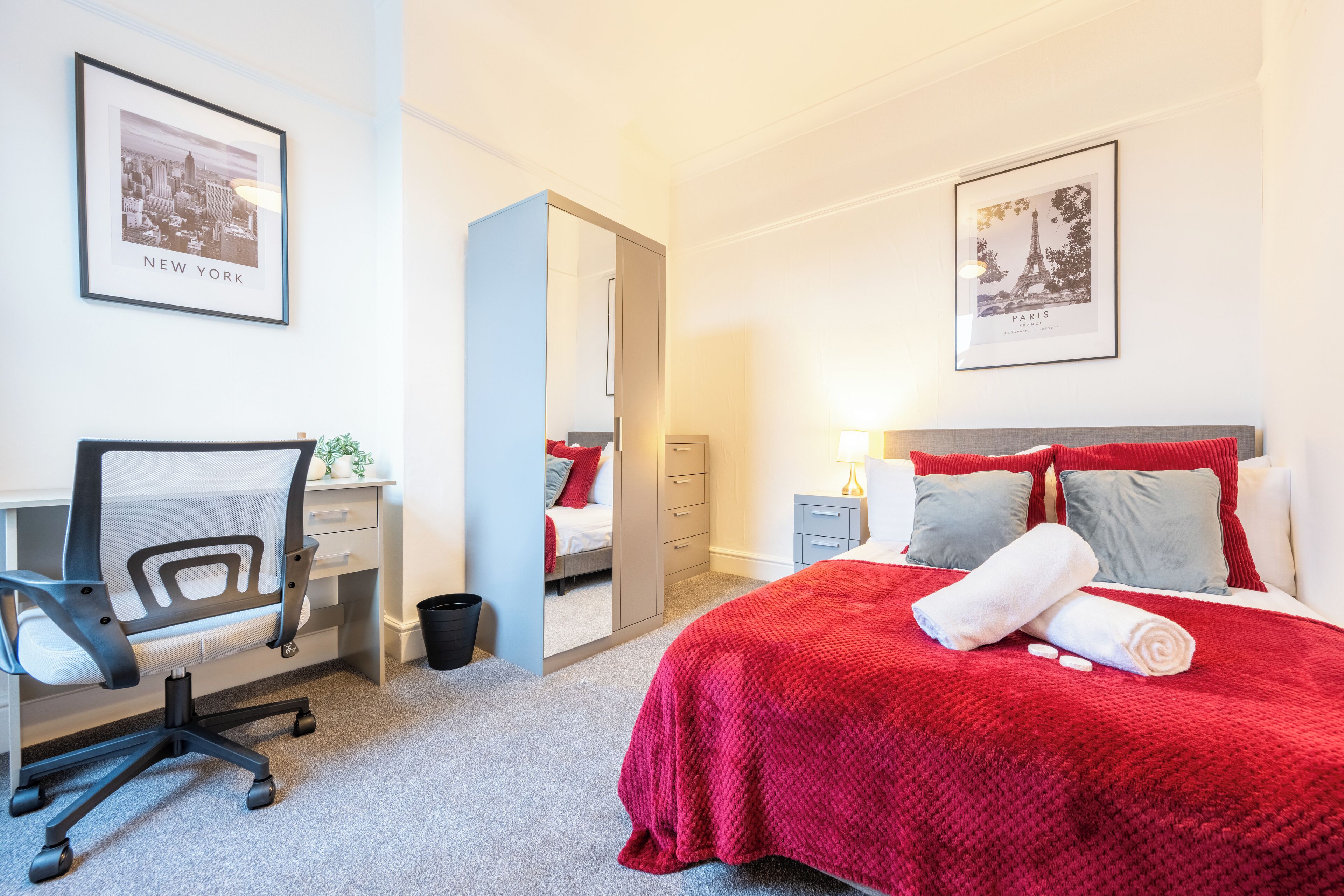 A hotel room with a bed, a desk, and a chair. There are framed pictures on the wall, a mirror, and a red blanket on the bed.