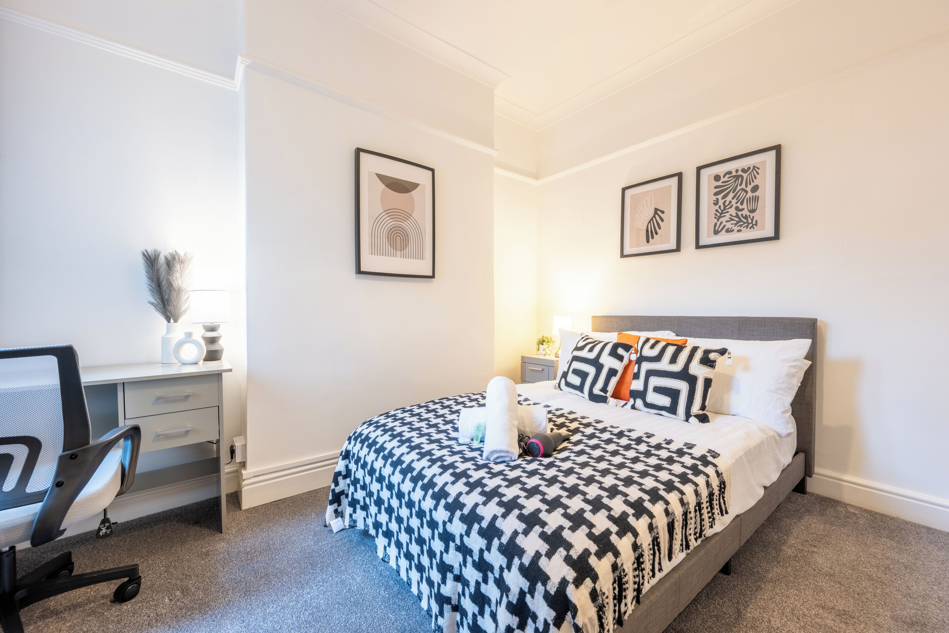 Un dormitorio ordenado con cama, escritorio y silla. Hay dos obras de arte enmarcadas en la pared.