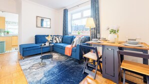 Ein modernes Wohnzimmer mit einem blauen Sofa, einem kleinen Esstisch und einem durch eine offene Tür sichtbaren Küchenbereich.