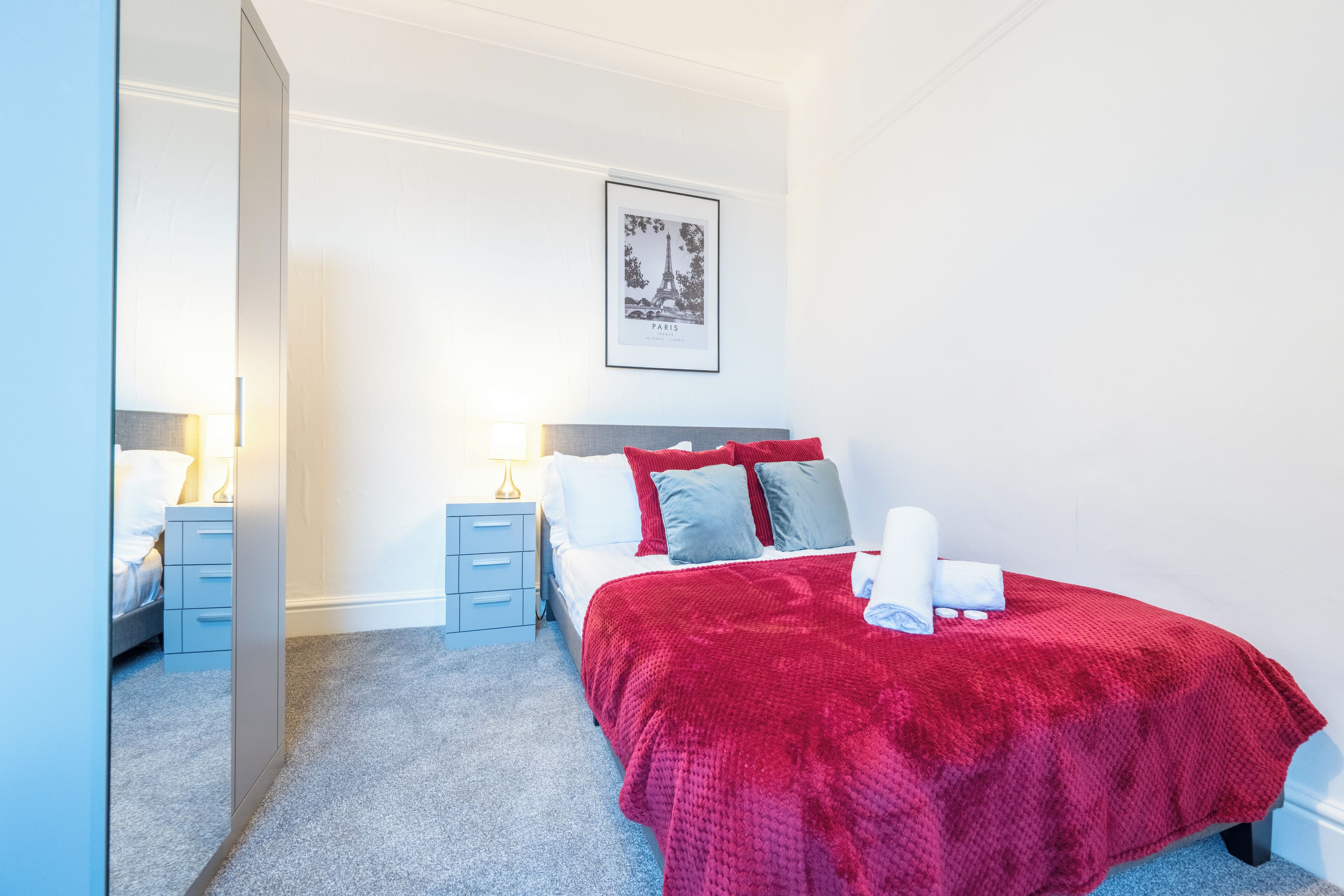 A bedroom with a bed, a nightstand, a lamp, a framed picture on the wall, and a carpeted floor.