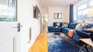 Ein modernes Wohnzimmer mit einem blauen Sofa, Holzboden, einem großen Fenster mit Vorhängen und einem an der Wand montierten Fernseher.