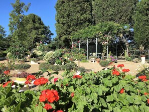 Ein Garten mit verschiedenen Pflanzen und Blumen, eine Pergola mit Sitzgelegenheiten und ein eingezäunter Bereich mit Tischen und Stühlen.