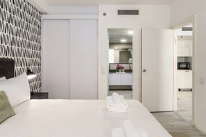 A hotel room with a bed, a nightstand, a wall-mounted lamp, a bathroom with a mirror, and a kitchenette in the background. - Beachfront 1BR • Balcony Views by SeaNRent (Tel-Aviv)
