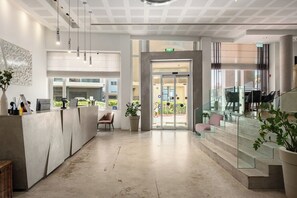 A modern hotel lobby with a reception desk, seating area, and a staircase leading to an upper level. - Beachfront 1BR • Balcony Views by SeaNRent (Tel-Aviv)