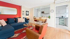 A modern living room with a blue sofa, a coffee table, and a red rug. There is a dining area with a wooden table and chairs. The kitchen is visible with white cabinets and a clock on the wall. - GuestReady - Santa Catarina Skyflat (Porto)