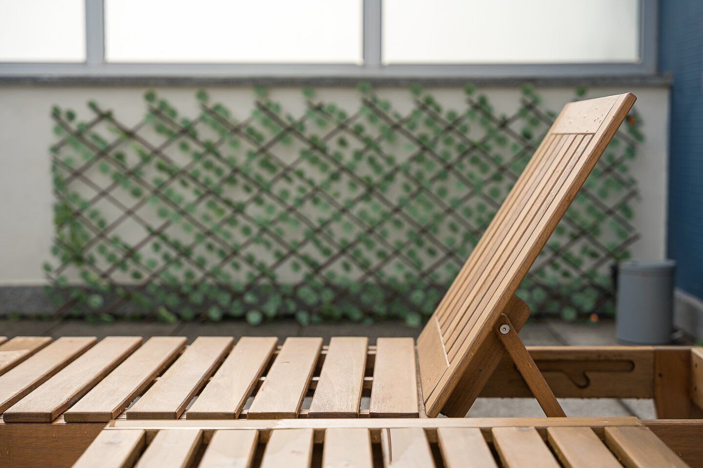 A wooden lounge chair with a backrest, positioned in front of a lattice fence with green foliage.