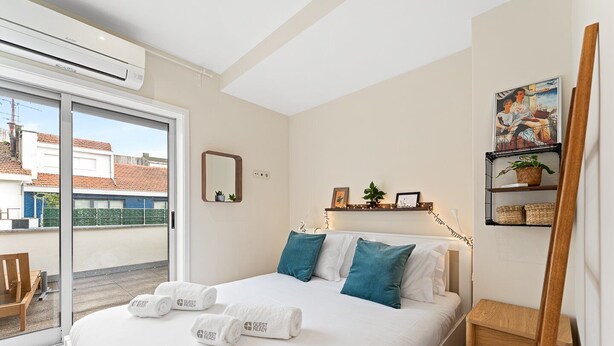 A hotel room with a bed, a chair, a small table, a shelf with plants, and a painting on the wall. - GuestReady - Santa Catarina Skyflat (Porto)