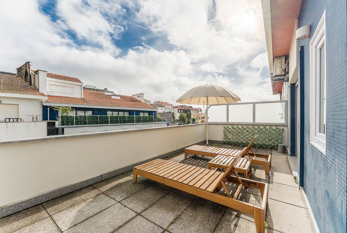 A rooftop terrace with wooden loungers, a large umbrella, and a view of the cityscape.