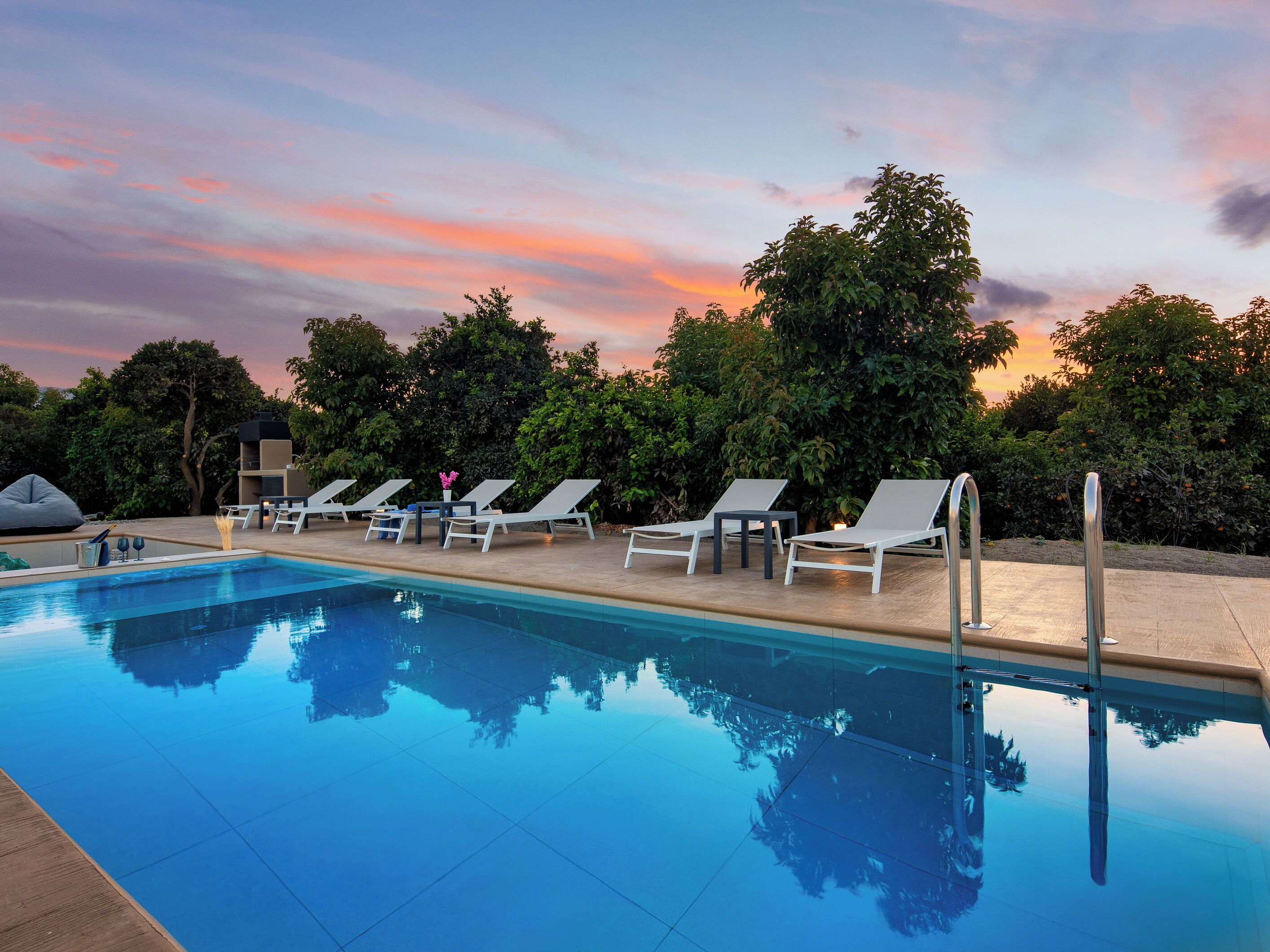 A swimming pool with lounge chairs and a table, surrounded by trees and a sunset sky.