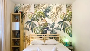 Ein Hotelzimmer mit einem Bett, einem Holz-Kopfteil, einem Nachttisch mit Lampe, einem Regal mit Dekoration und einer Wand mit tropischem Blattmuster.