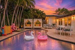 A pool with swan and flamingo floats, a cabana, outdoor furniture, and a building with lights. - Designer 4BR Villa • Pool • 30 min to WC Stadium (Fort Lauderdale)