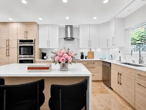A modern kitchen with white cabinetry, a central island, a stainless steel range hood, and a window with blinds. - Designer 4BR Villa • Pool • 30 min to WC Stadium (Fort Lauderdale)