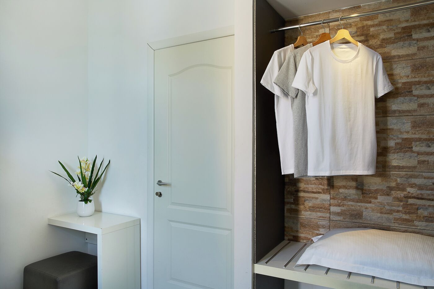 Une pièce avec une porte blanche, une petite table blanche ornée d’une plante, et un lit avec un oreiller blanc adossé à un mur de briques.