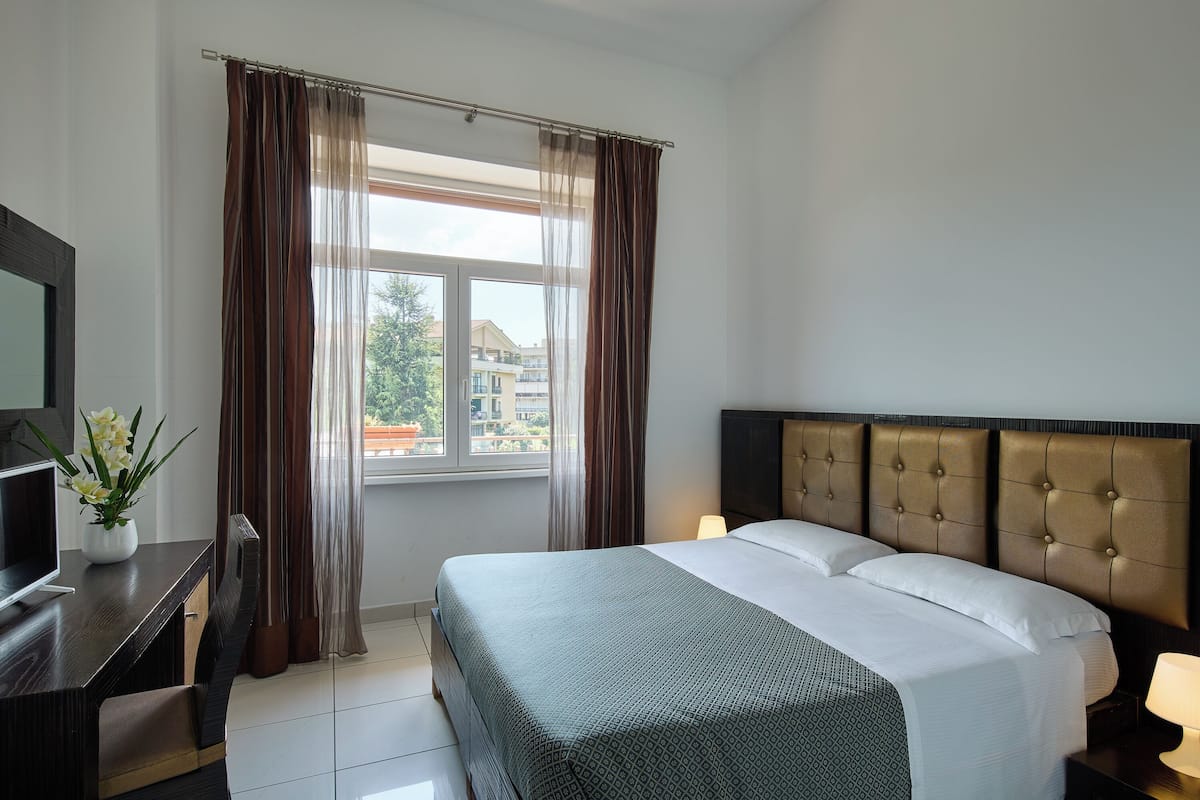 A hotel room with a bed, a desk with a TV, a chair, and a window with curtains.