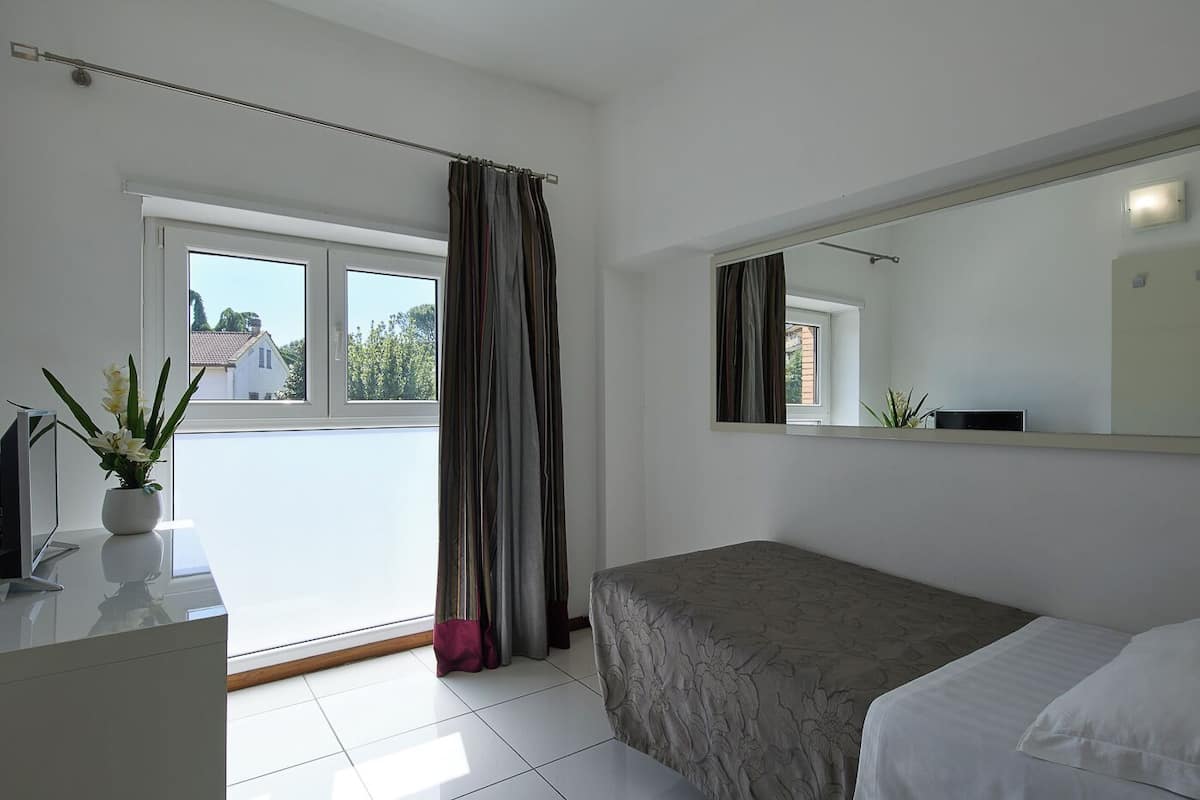 A bedroom with a bed, a bedside table, a mirror, a window with curtains, and a potted plant.