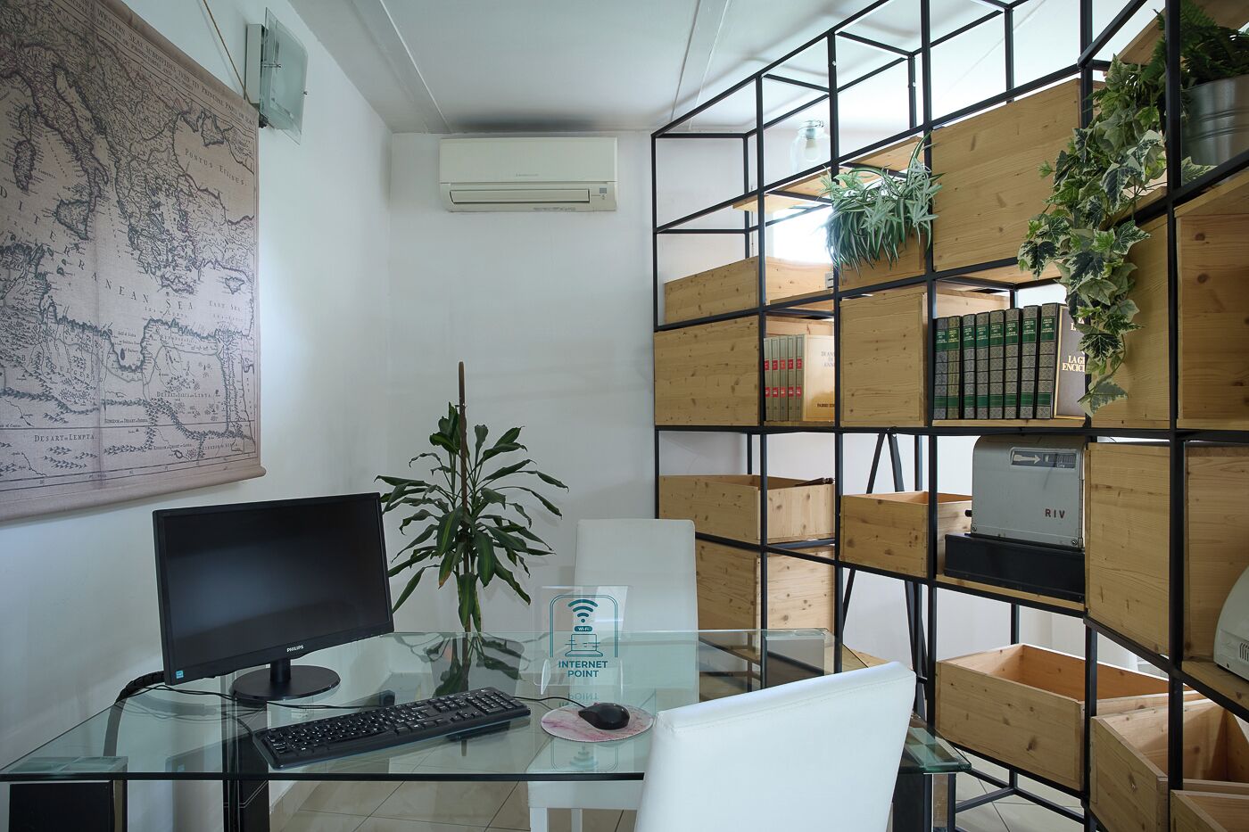 A modern office space with a glass desk, a computer monitor, keyboard, and a plant. There is a map on the wall and a bookshelf with books and plants.