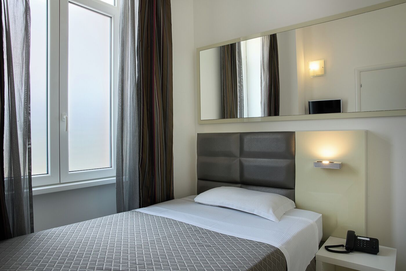 Una habitación de hotel con cama, teléfono junto a la cama, espejo y ventana con cortinas.