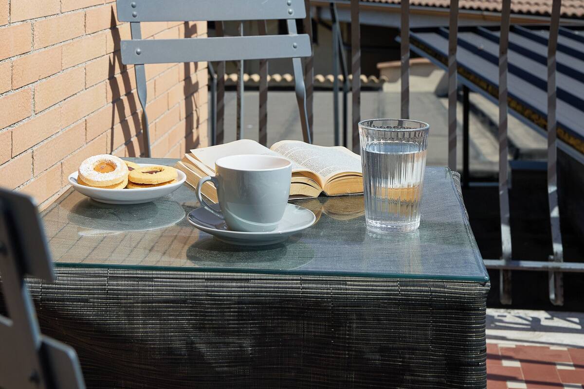 A table with a glass of water, a cup, and a plate with cookies on a balcony with a brick wall and a tiled roof.