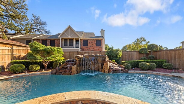 A backyard with a swimming pool, a waterfall feature, and a house with a brick facade and a gabled roof. - Luxury Pool Home w/ Outdoor Lounge Near Stadium (Irving)