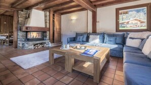 Un salón acogedor con chimenea de piedra, vigas de madera, un sofá azul y vista a una cabaña de madera a través de un amplio ventanal.