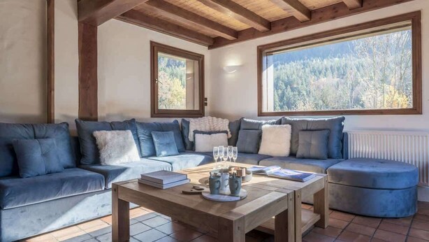 Una sala de estar moderna con techo de madera, un sofá azul, una mesa de centro de madera y vistas a un bosque a trav és de grandes ventanales.