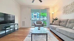 Ein Wohnzimmer mit Flachbildfernseher, Couch, Couchtisch, Deckenventilator und Blick auf ein Gebäude durch eine Glastür.