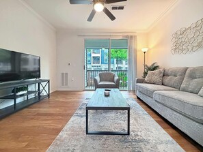 Ein Wohnzimmer mit Flachbildfernseher, Couch, Couchtisch, Deckenventilator und Blick auf ein Gebäude durch eine Glastür.