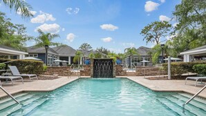 A swimming pool with a waterfall feature, surrounded by lounge chairs and umbrellas, with a clear sky above. - Peaceful 2 BR with Private Garage, Prime North Tampa (Lutz)