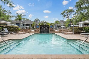 A swimming pool with a waterfall feature, surrounded by lounge chairs and umbrellas, with a clear sky above. - Peaceful 2 BR with Private Garage, Prime North Tampa (Lutz)