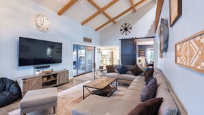 A modern living room with a large flat-screen TV, wooden beams on the ceiling, and a cozy seating area with a sofa and ottoman.