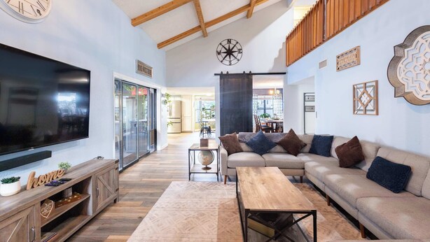 A spacious living room with a large flat-screen TV, wooden furniture, a sofa with cushions, a coffee table, and a wooden ceiling with exposed beams.