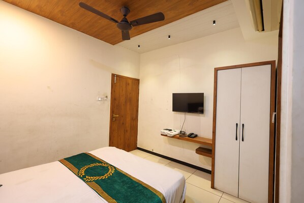 Quarto de hotel com cama, ventoinha no teto, televisão, guarda-roupa e uma pequena estante com objetos.