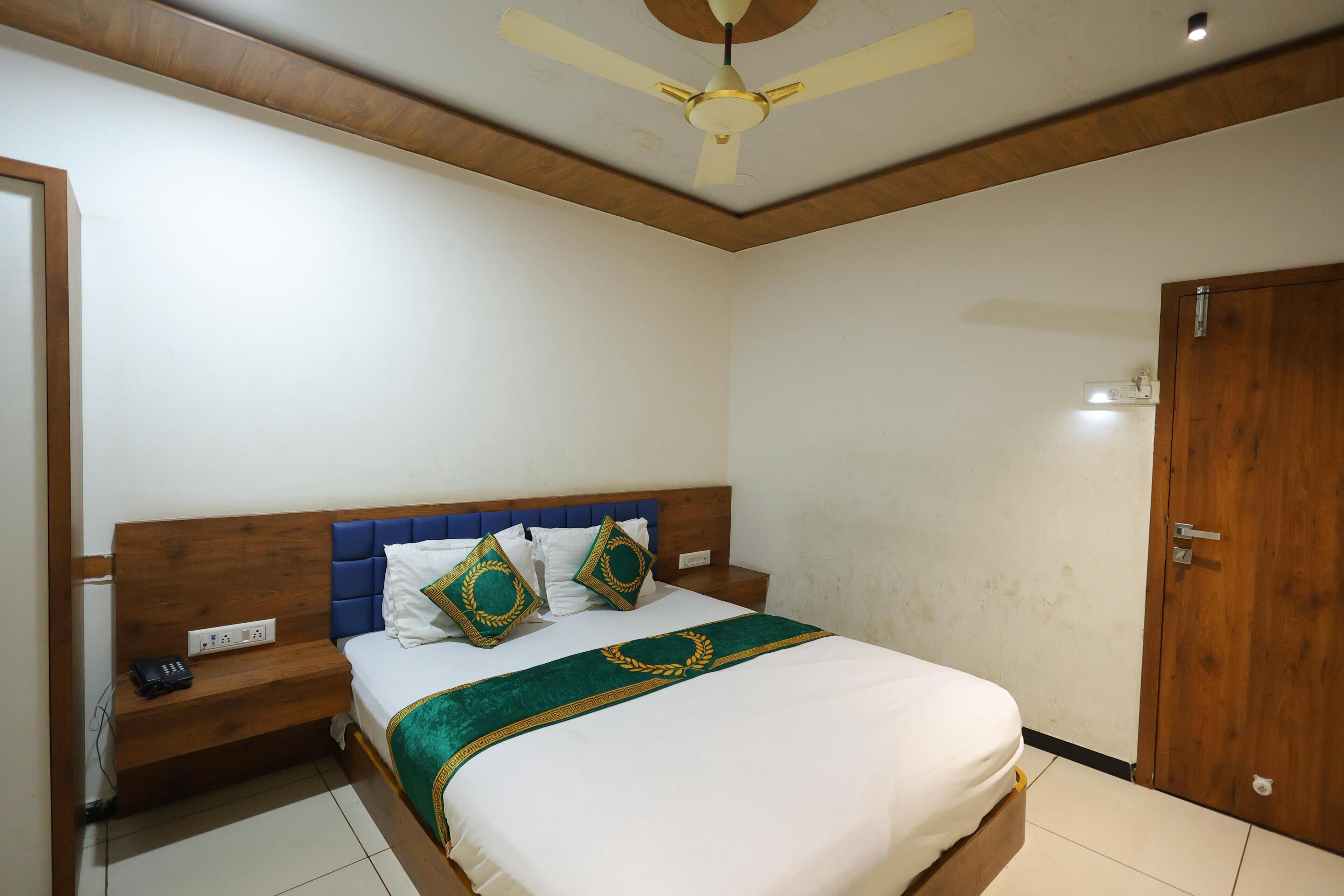 A bedroom with a wooden headboard, a bed with white and green bedding, a nightstand, a power outlet, and a ceiling fan.