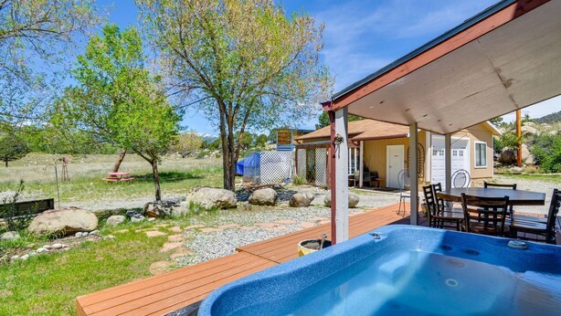 Un jacuzzi en una terraza de madera con mobiliario exterior, rodeado de un jardín ajardinado con árboles y rocas.