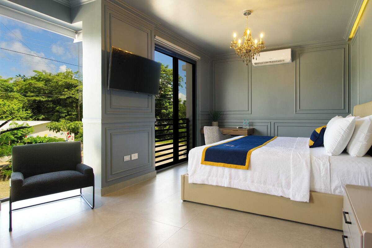 A bedroom with a bed, a chair, a TV, and a window with a view of greenery.