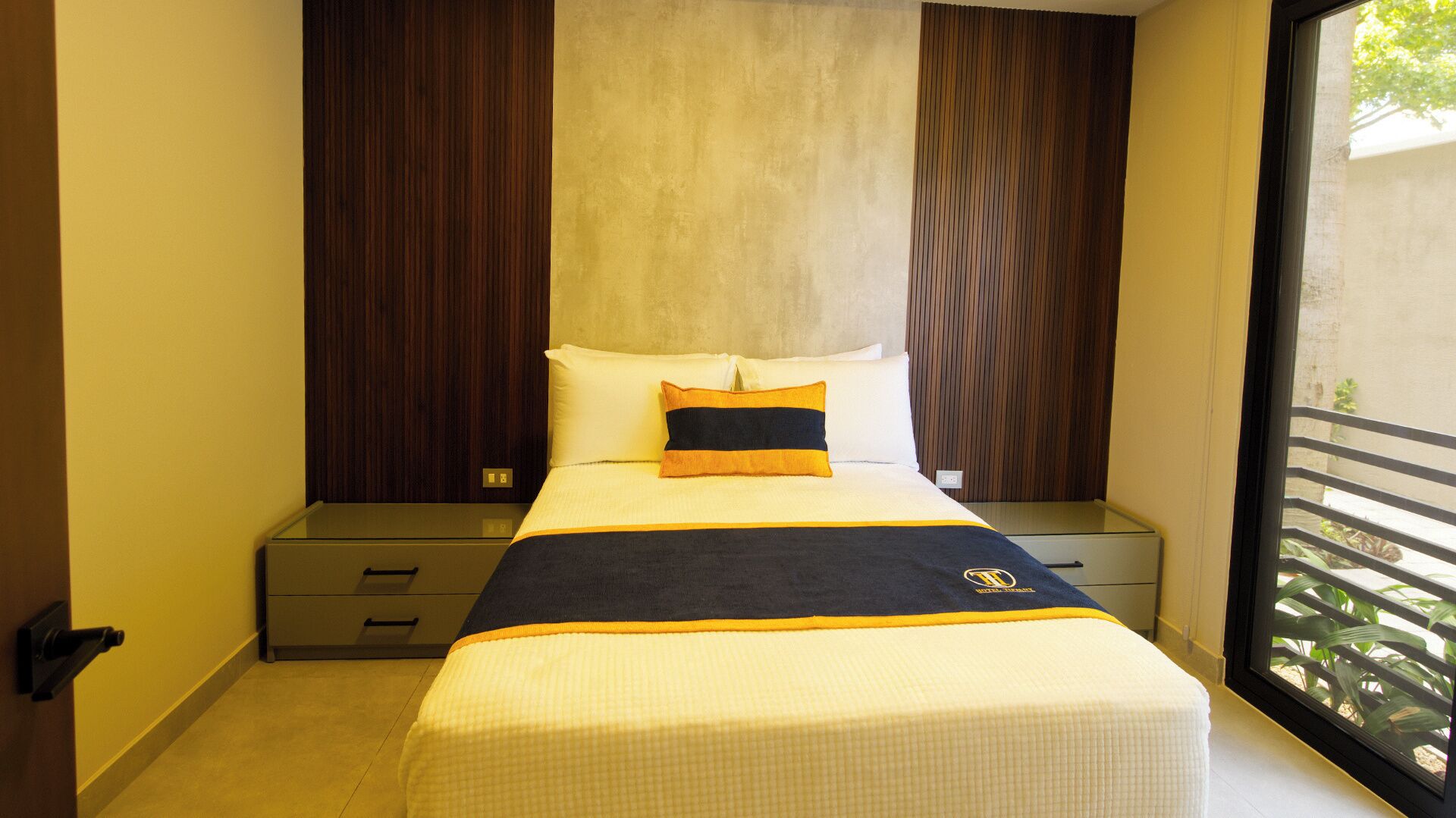 A hotel room with a bed, a nightstand, a drawer unit, and a window with a view of greenery.