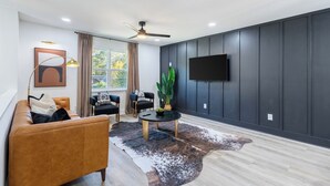 Un salon moderne comprenant un canapé, deux fauteuils, une table basse, une grande plante, un téléviseur fixé au mur et un ventilateur de plafond.