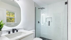 Un bany modern amb plat de dutxa envoltat de vidre, un lavabo blanc amb taulell de marbre i un mirall gran.