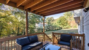 Une terrasse extérieure avec deux canapés et une table, entourée d’une rambarde en bois, donnant sur une cour avec des arbres et une maison en arrière-plan.