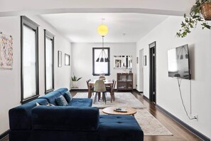 Een moderne woonkamer met een blauwe bank, een eethoek met een tafel en stoelen, en een aan de muur bevestigde televisie.