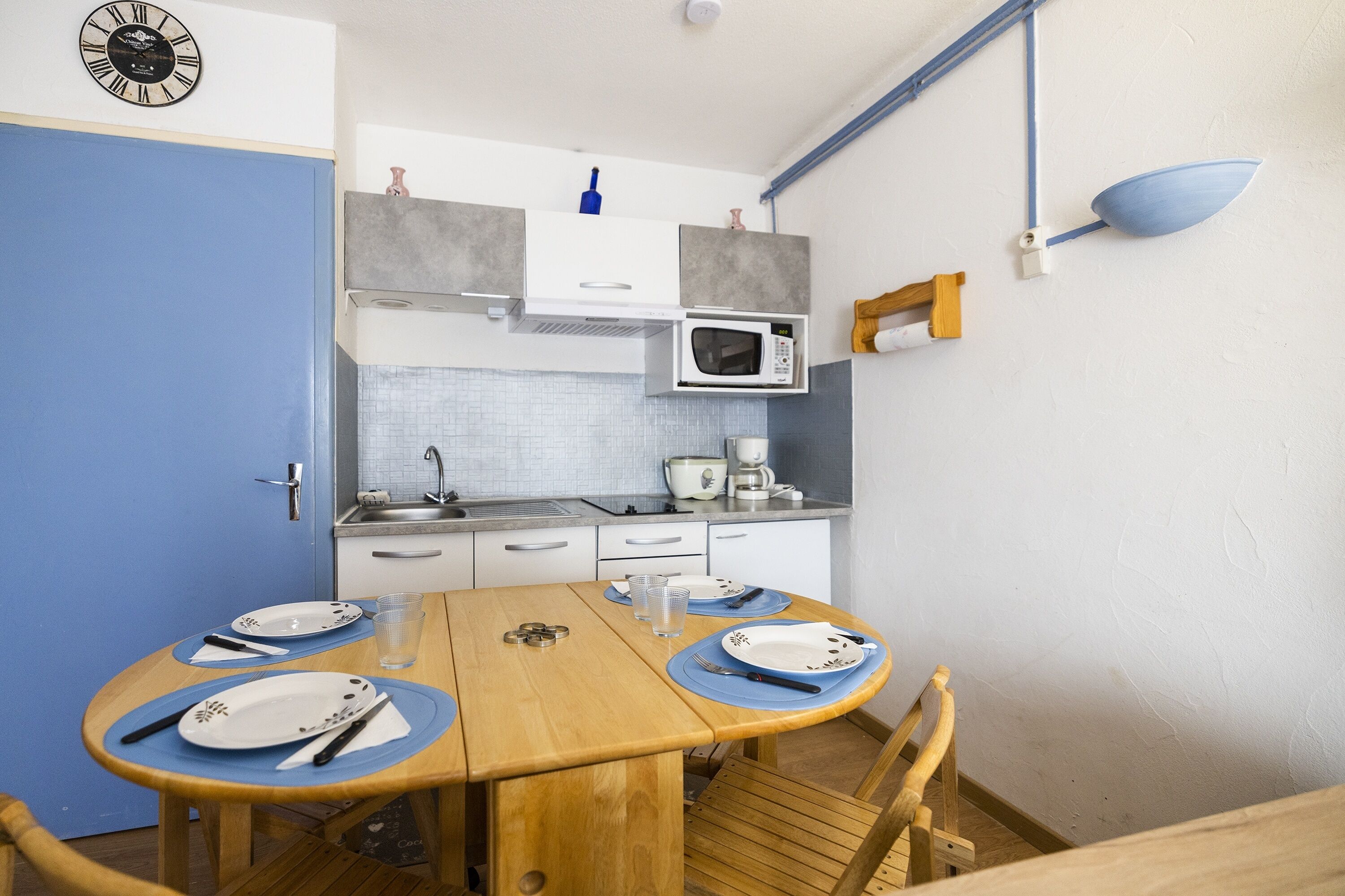 A compact kitchen with a microwave, sink, and cabinets. There is a dining table with plates and cutlery.