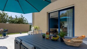 Um pátio externo com mesa, cadeiras, um sofá de vime, um guarda-sol grande e uma porta de vidro que dá para um espaço interno.