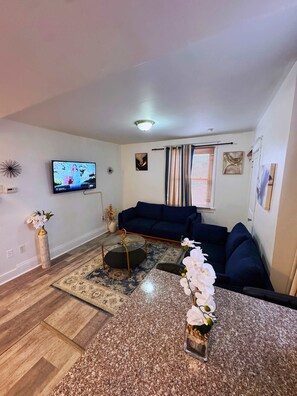 Ein Wohnzimmer mit einem dunkelblauen Sofa, einem Glastisch, einem an der Wand montierten Fernseher und einer dekorativen Vase mit Blumen.