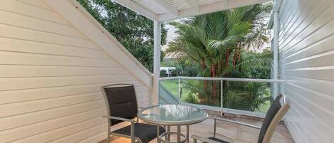 A small balcony with a glass table and two chairs, surrounded by white walls and a view of palm trees.